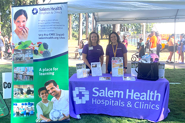 Two Salem Health CHEC staffers standing behind info table at community event.