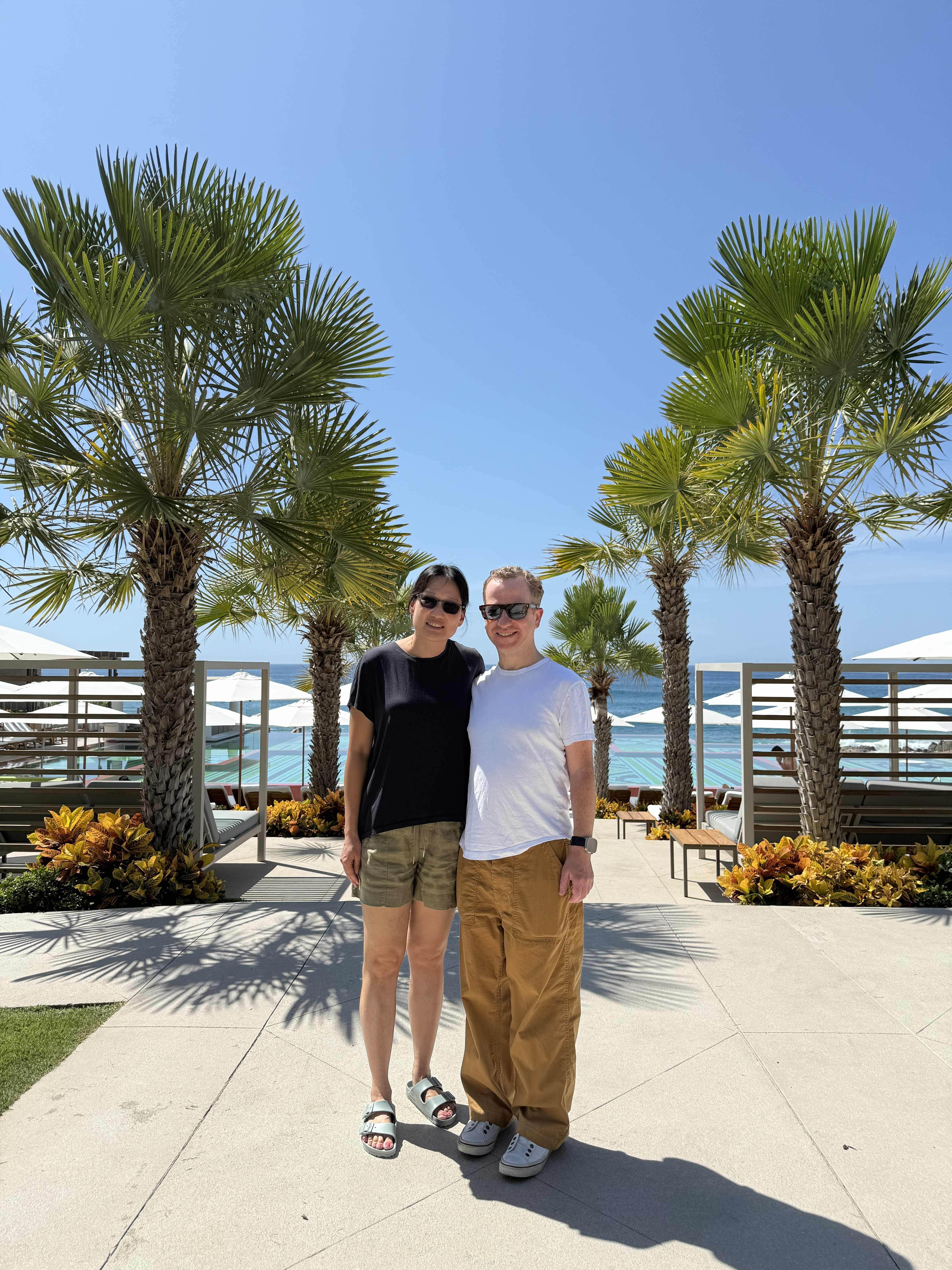 Dr. Egalka and his wife stand in front of palm trees and the ocean.