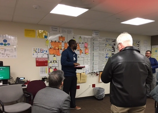 Four men in a conference room looking at a wall of charts and graphs.