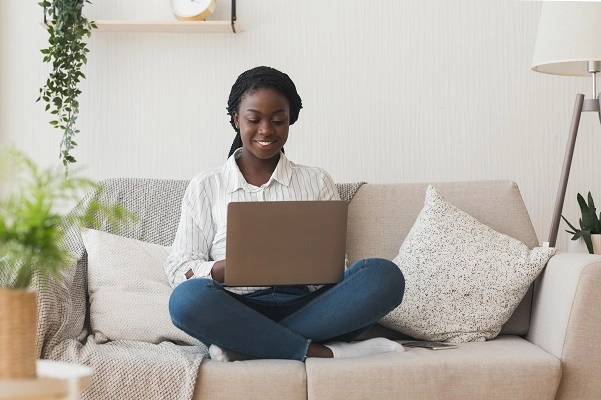A 20-something African American woman works on a laptop, cross-legged on a couch.