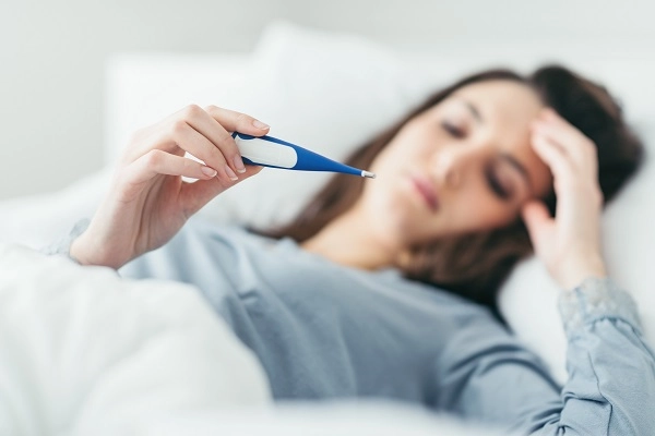 A young woman in bed looks checks a thermometer.