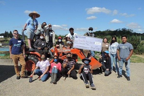 Staff of Marion-Polk Food Share pose on a tractor outside their warehouse with a novelty check from Salem Health