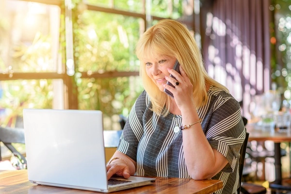 An older blonde woman talks on the phone while typing on a laptop.